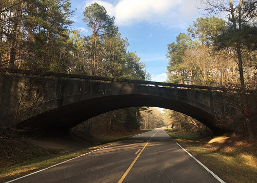 Stone bridge over road that disappears into trees ahead. Some sunlight coming through clouds onto the road.