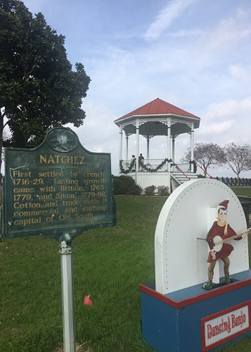 Natchez sign at left, gazebo at center, and figure of man playing a banjo at lower right, all in a park. Blue sky with clouds above.