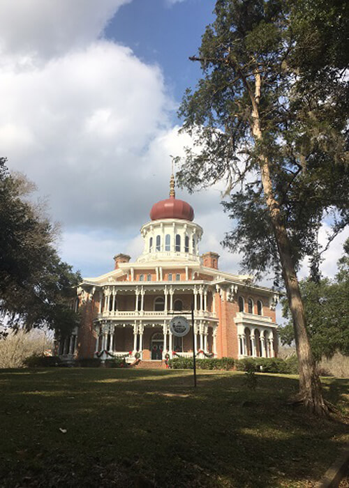 Ornamental octagon house with red sphere perched on top of white circular tower. Blue sky with some thick white clouds.