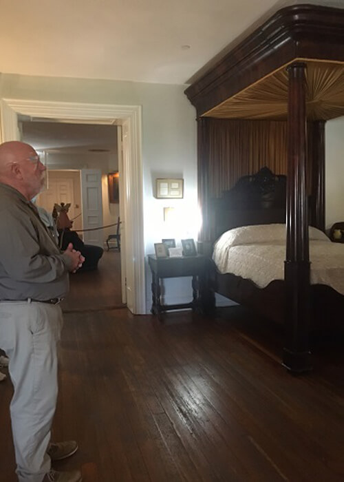 Room inside the antebellum Longwood Mansion, with wooden floors, bed at right, and tour guide at left explaining the history.