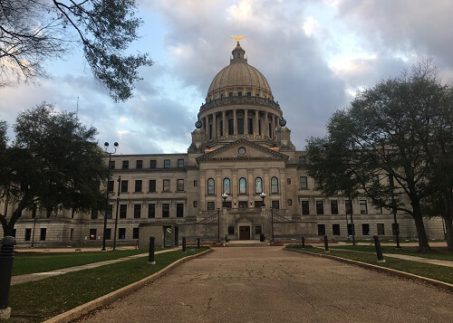 Path leading to front Mississippi State Capitol building, blue sky above with some thick clouds. Trees on both sides of the pathway.