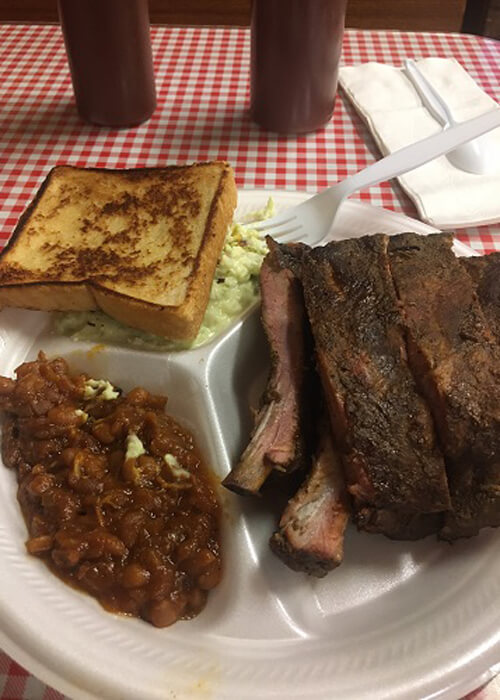 Plate of barbecue ribs, baked beans and cheese bread on red and white checkered table cloth.