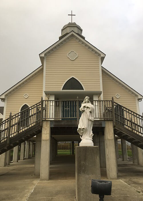 Beige-colored church on cement stilts, statue of biblical figure in front. Hazy grey sky above.