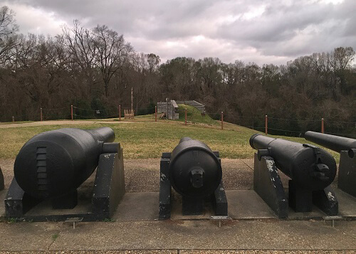 Three big cannons facing a green field with wall of trees in the distance, part of the Vicksburg National Military Civil War site. Cloudy grey sky above.