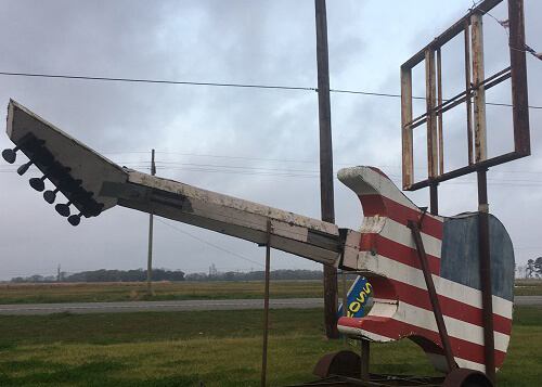 Massive guitar painted red white and blue as a roadside amusement. Grey clouded sky above.