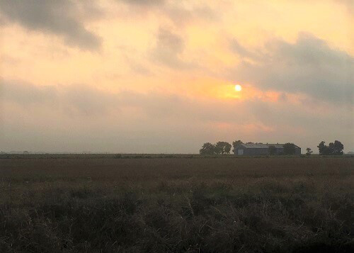 Sun coming through thick orange clouds above an open field with a farm in the distance.