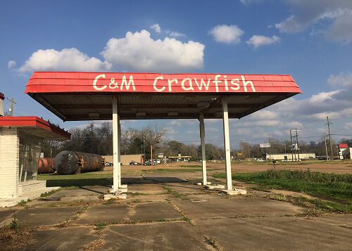 Red gas station canopy with "C&M Crawfish" painted on the side. Gas station is abandoned with some weeds growing from the cracks in pavement.