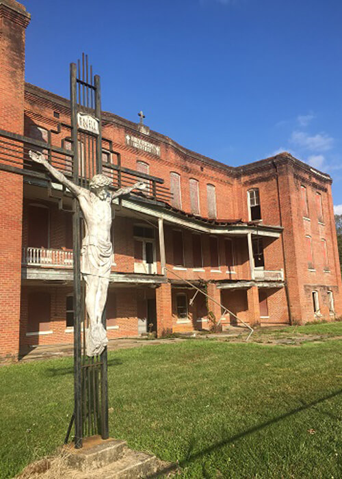 Three story abandoned brick building with the windows boarded up or missing. Figure of Christ hanging from cross in front. Clear blue sky.