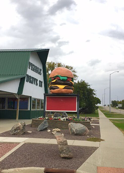 Green roofed restaurant with sign saying "Pebble Drive-In".  Four-foot tall giant cheeseburger in front of restaurant.