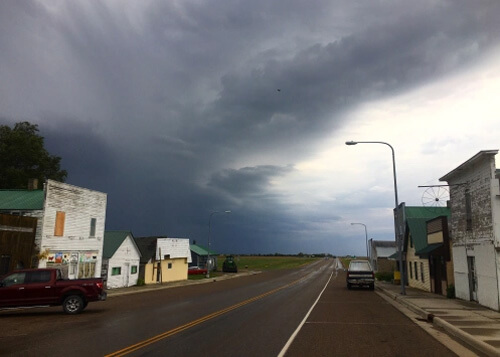 Road disappearing into dark storm cloud formation, a few white single story buildings on both sides of road with weathered paint.