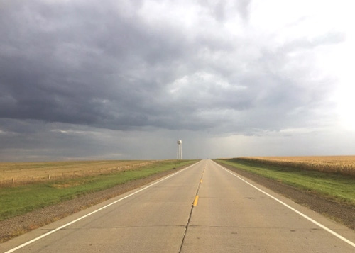 Road disappearing straight ahead into the horizon underneath storm clouds and with corn fields on both sides.  Water tower in distance.