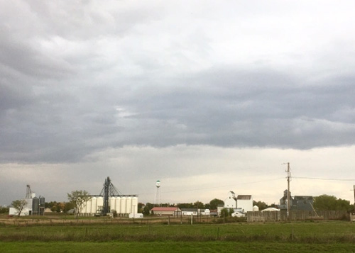 Small town with grain silos and water tower at bottom of photo, storm clouds above.