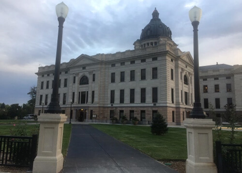 Four-story state capitol building of South Dakota with green dome protruding from center of cement building.  Two lamp posts on both sides of the narrow road leading to building.  Cloudy sky above.