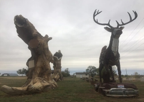 Huge deer statue at right on top of old Buick car, log structure at left.  Grey skies.