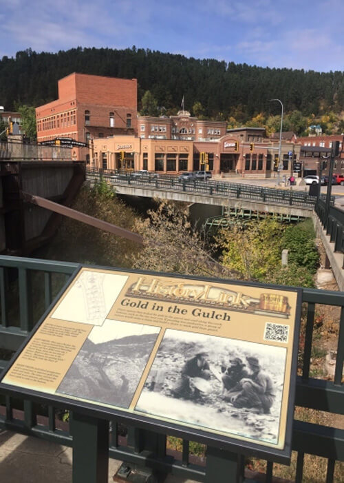 A few brick buildings across a river with a sign at bottom that reads "Fold in the Gulch."  Forest trees behind buildings and blue sky above.