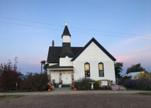 Plain white house with dark shingled roof with some flowers in front, blue and pink sky above at sunrise.