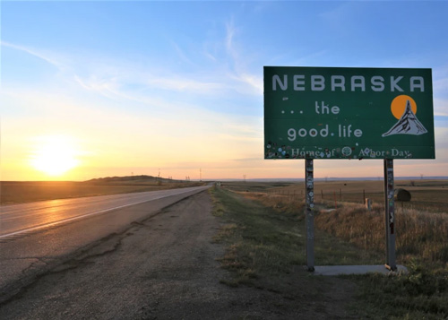 Sign at right reading "Nebraska, the good life" with road at left and sun rising above the road.  Blue sky above.