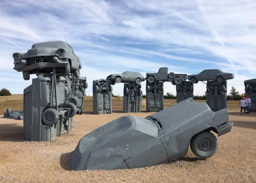 Semicircle of grey painted cars as part of pop art monument, blue sky above with some clouds.