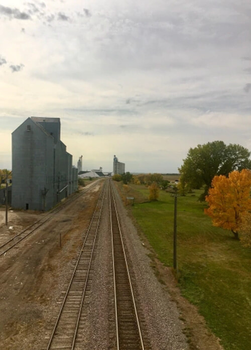 Railroad track extending into the horizon, grain silos at left and a few trees at right.  Cloudy grey sky above.