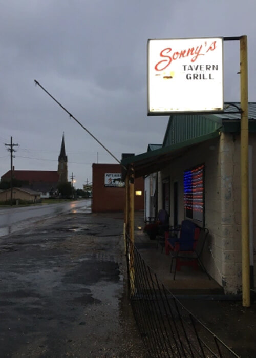 Sunrise photo with dark sky and a few buildings and a church steeple, and a bright lit sign at upper right that reads "Sonny's Tavern Grill."