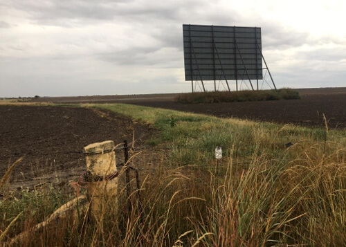 Abandoned drive-in theatre screen in a dirt field, grey cloudy skies above.