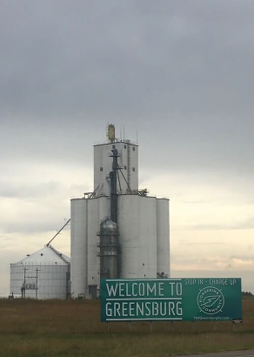 Grain silo and sign in front saying "welcome to greensburg" with cloudy skies behind.