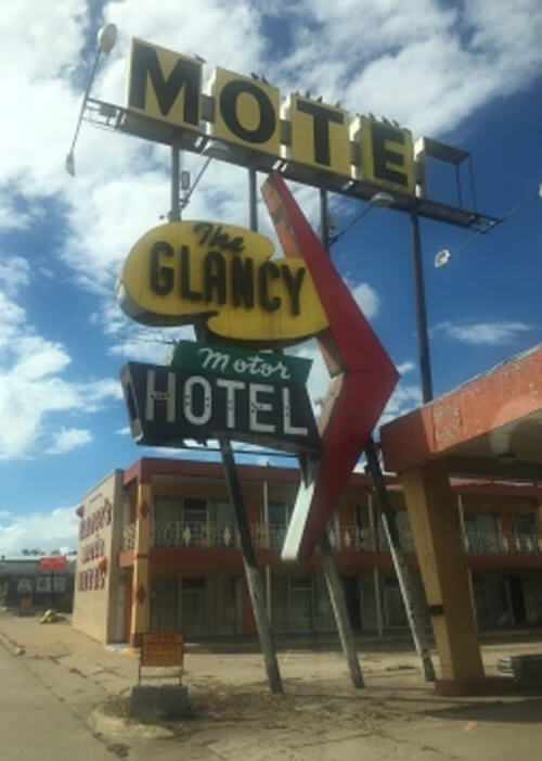 1960's style motel sign with missing letters saying "The Clancy Motor Motel" in front of abandoned motel with blue sky and scattered clouds behind.