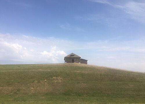 Sparse wooden house sitting by itself on a green prairie hill, blue sky a few clouds above.