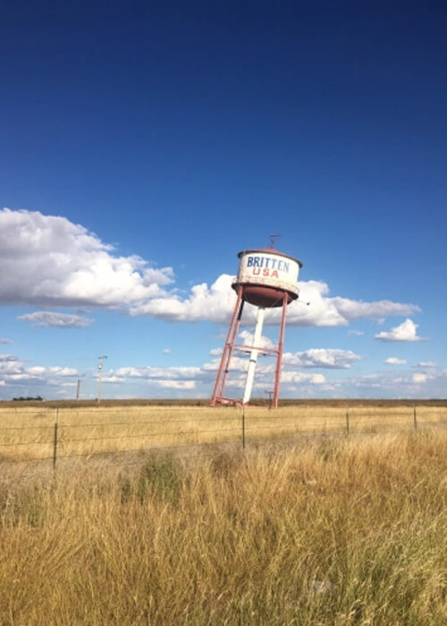 Leaning water tower in grass field that says "Britten USA" with blue sky and a few scattered clouds behind it.