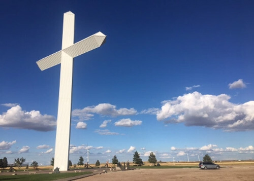 Multi-story high cross at left, and van at right looking tiny in comparison.  Blue sky and scattered clouds behind the cross.