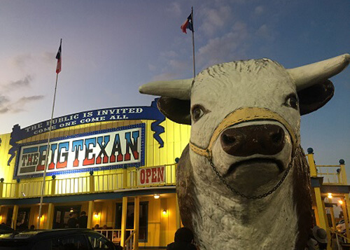 Cow head statue at right and yellow restaurant at left with sign that reads "The Big Texan" at dusk.