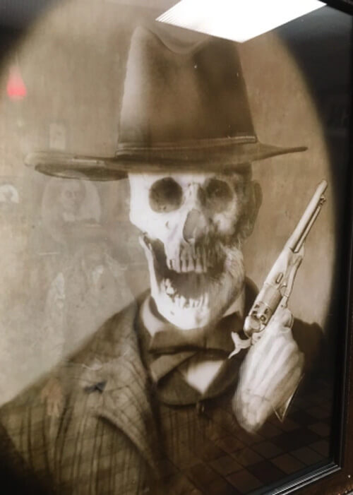 Black and white portrait picture of a skull with hat and holding gun.