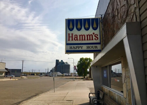 Sign above bar saying "Hamm's Happy Hour", street at left.  A grain silo in distance, blue sky above with a few clouds.