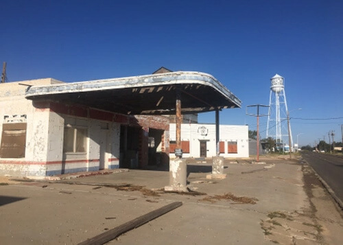 Abandoned gas station at left with water tower in distance that says "home town of Waylon Jennings."