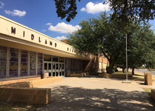 Front of Midland High School, light tan single story building that says "Midland High" at left and some trees at right.