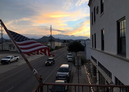 Looking down on street from second story of hotel at sundown, a car driving down road below and American flag waving from balcony.  Orange clouds in distance.