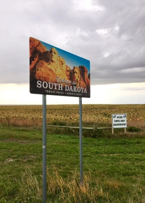 Sign reading "welcome to North Dakota" with sunflower field behind it, cloudy grey skies above.