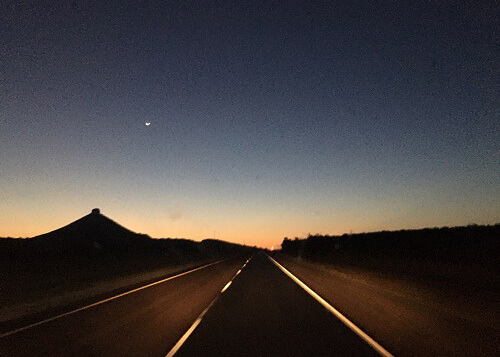 Orange peering over the horizon at sunrise with road disappearing over a slight hill.  Pointed hill at left of road.