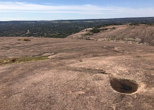 Red-brown dome rock with a crater formation and green trees off in the distance.  Blue sky with some clouds above.