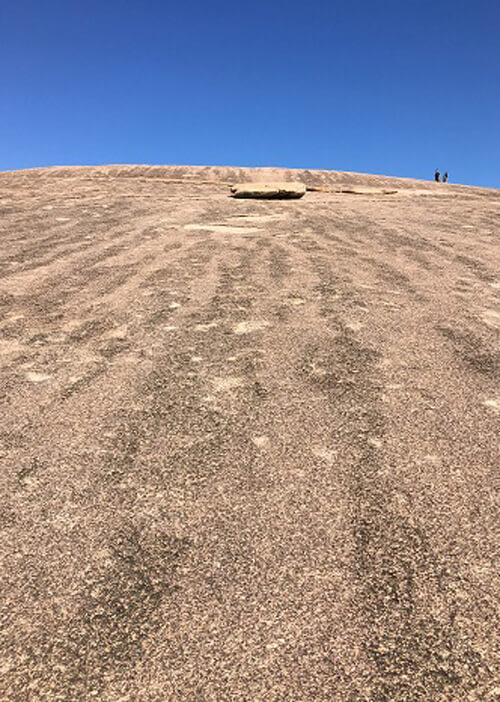 Looking up a dome shaped of a red-brown color, a few people far off in distance at top of rock.