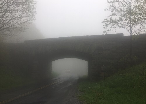 Road bridge made of stones with a couple leafless trees on either side on a very foggy morning.