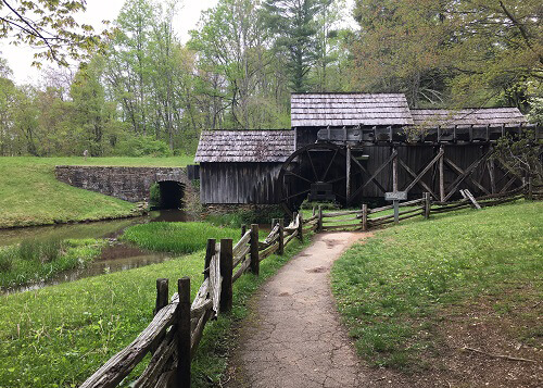 Walking path leading to a wooden mill built in 1910, green grass on both side of path, stream on the left.