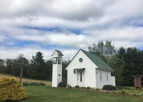 Small one-room white church with a big lawn and some trees in background, blue sky above covered in clouds.