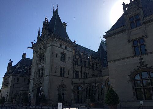 The front of Biltmore Estate, a massive Chateau-style home made of stone, blue sky above and sun peering over the roof. 