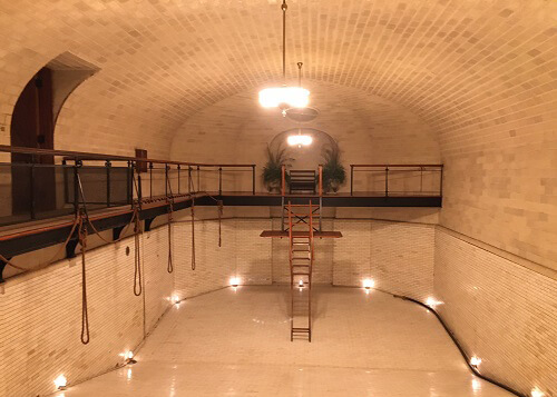 Indoor swimming pool inside Biltmore Estate, made of white stone and brick. Pool is empty with no water and lit up with lights.