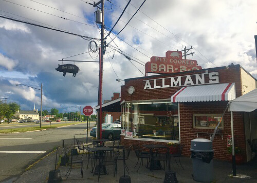 Small single-story brick barbeque restaurant with large front window and sign on building that says "Allman's". Blue sky covered in puffy white clouds.