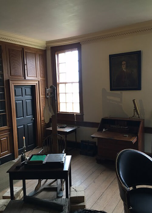 The den room inside George Washington's house, with a couple desks, chairs and a painting on the wall. Bright light coming through window.