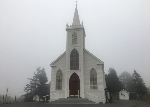 White clapboard covered single story wooden church with steeple and cross at top.  A few trees on the side and fog in background.