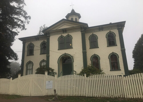 Looking up at two-story wooden schoolhouse, painted white with green trim. White picket fence in front, fog in the background.