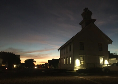 Sundown, near-dark photo of two story church at right and a few lights at the left.  Dark sky with a little orange at horizon.